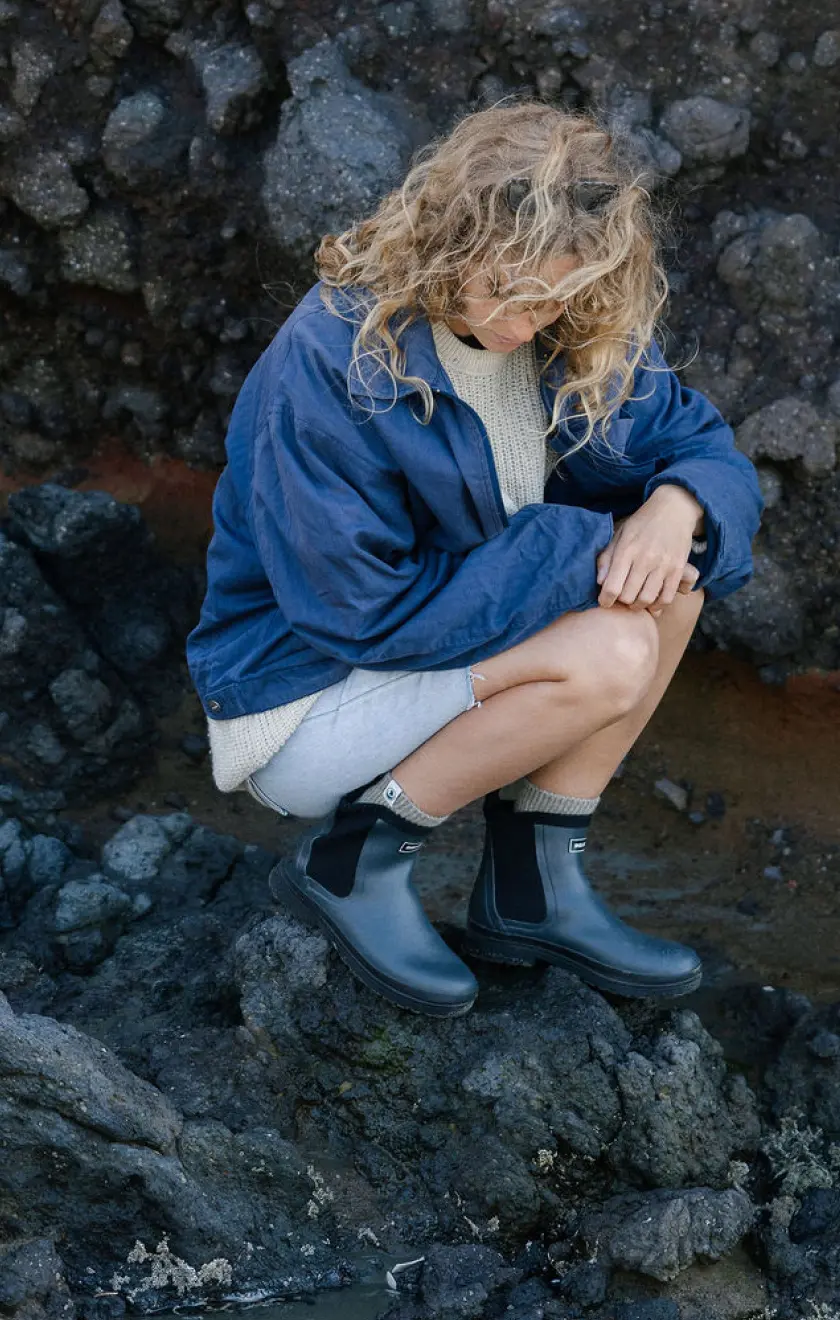 A person with curly blonde hair wearing a blue jacket, gray sweater, shorts, and black boots squats on rocky terrain, looking down at the ground.