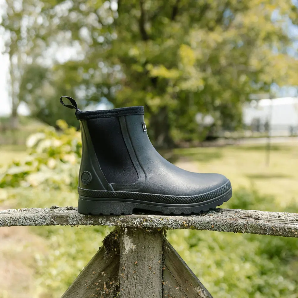 A single black rubber boot is balanced on a weathered wooden fence outdoors, with green trees and grass in the blurred background on a sunny day.