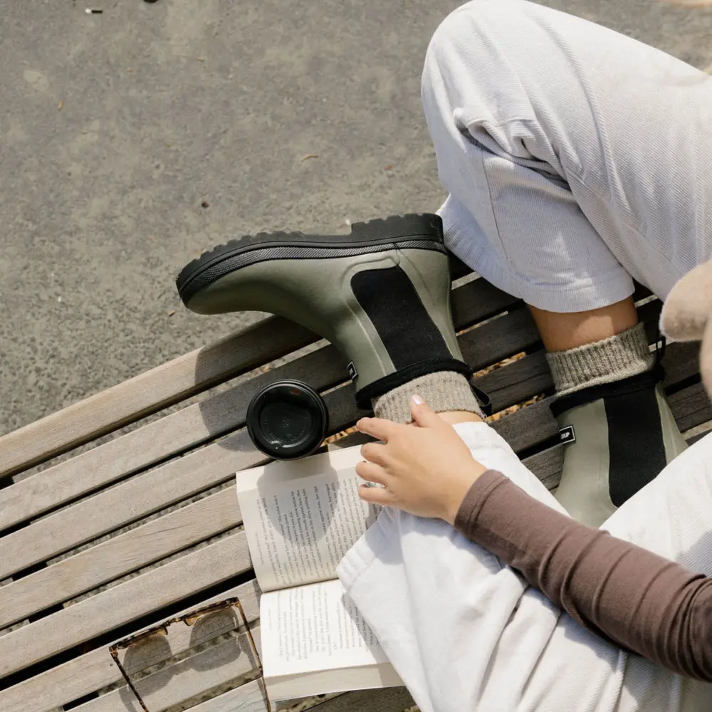 Person sitting on a wooden bench outdoors, wearing light pants, a brown long-sleeve top, and olive-green boots. They are reading a book with a takeaway coffee cup and glasses beside them.