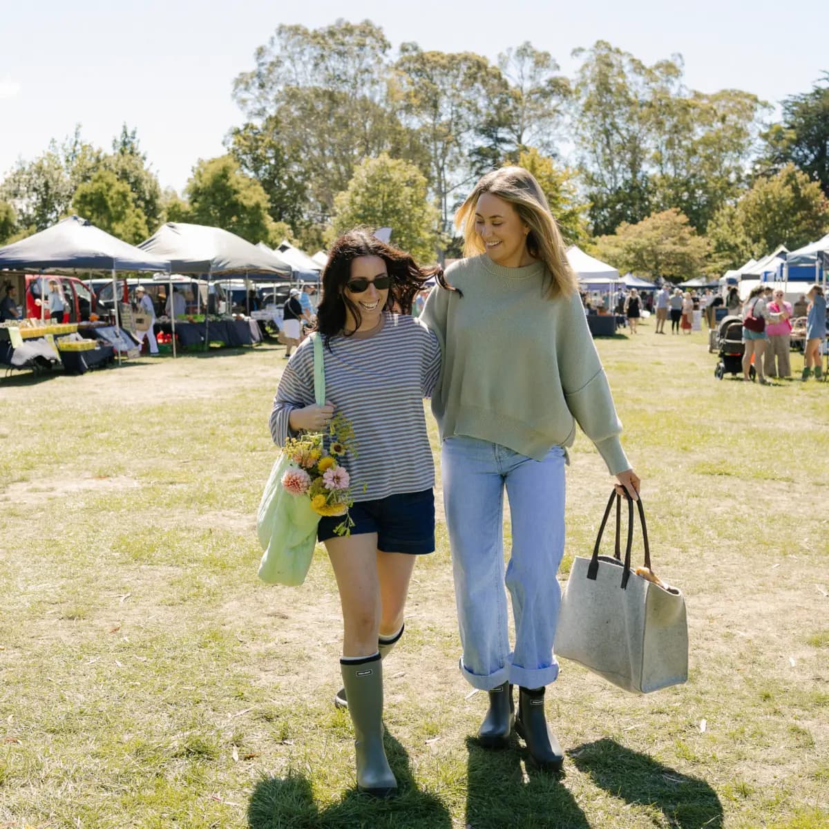 Two women walk together at an outdoor market, smiling and carrying bags. One holds flowers and wears shorts with rain boots; the other wears jeans and a sweater with ankle boots. Market stalls and trees are visible in the background.