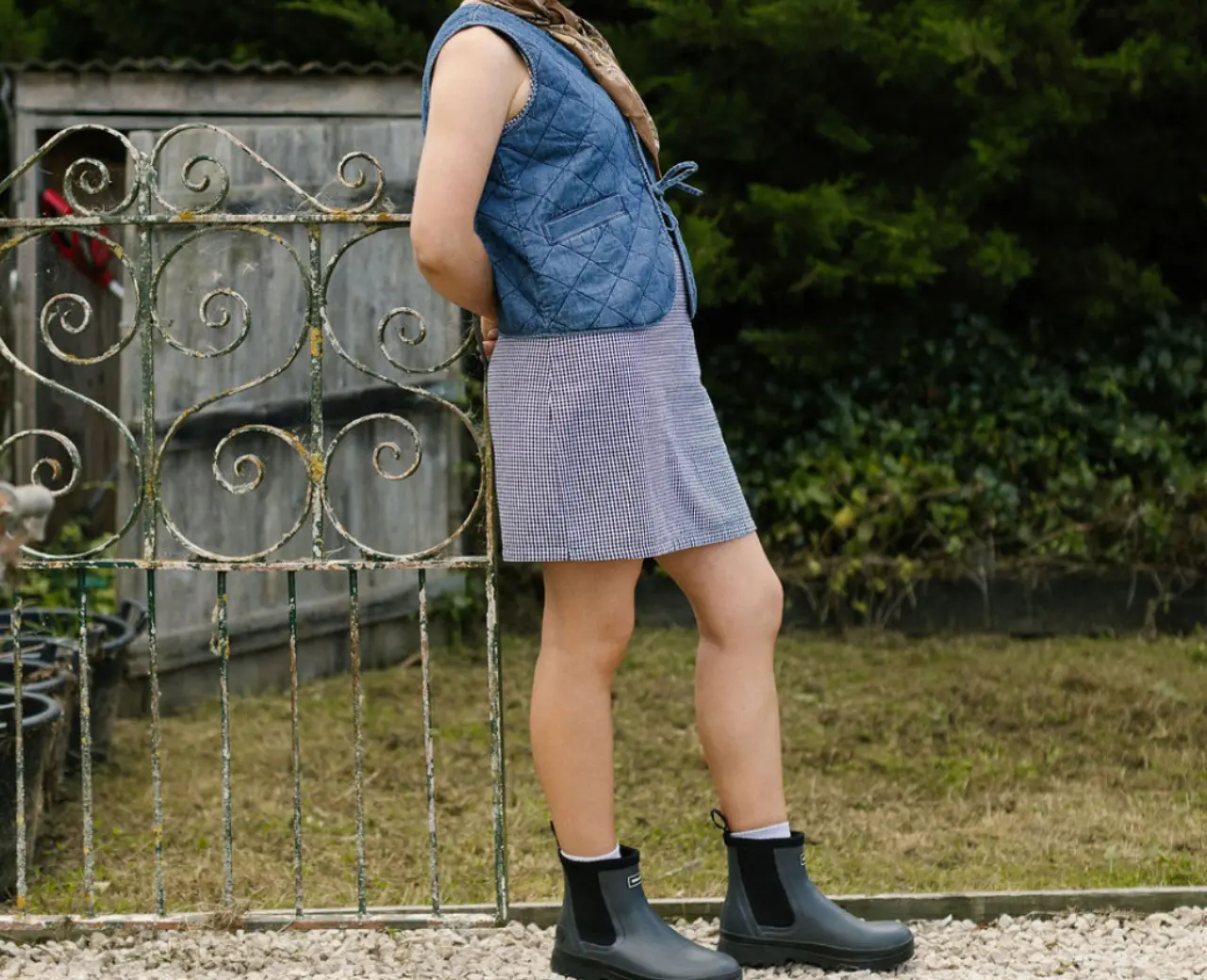 A person with long hair in a braid stands outdoors on gravel, wearing a blue quilted vest over a striped dress and black ankle boots. Green bushes and a rustic metal gate are in the background.