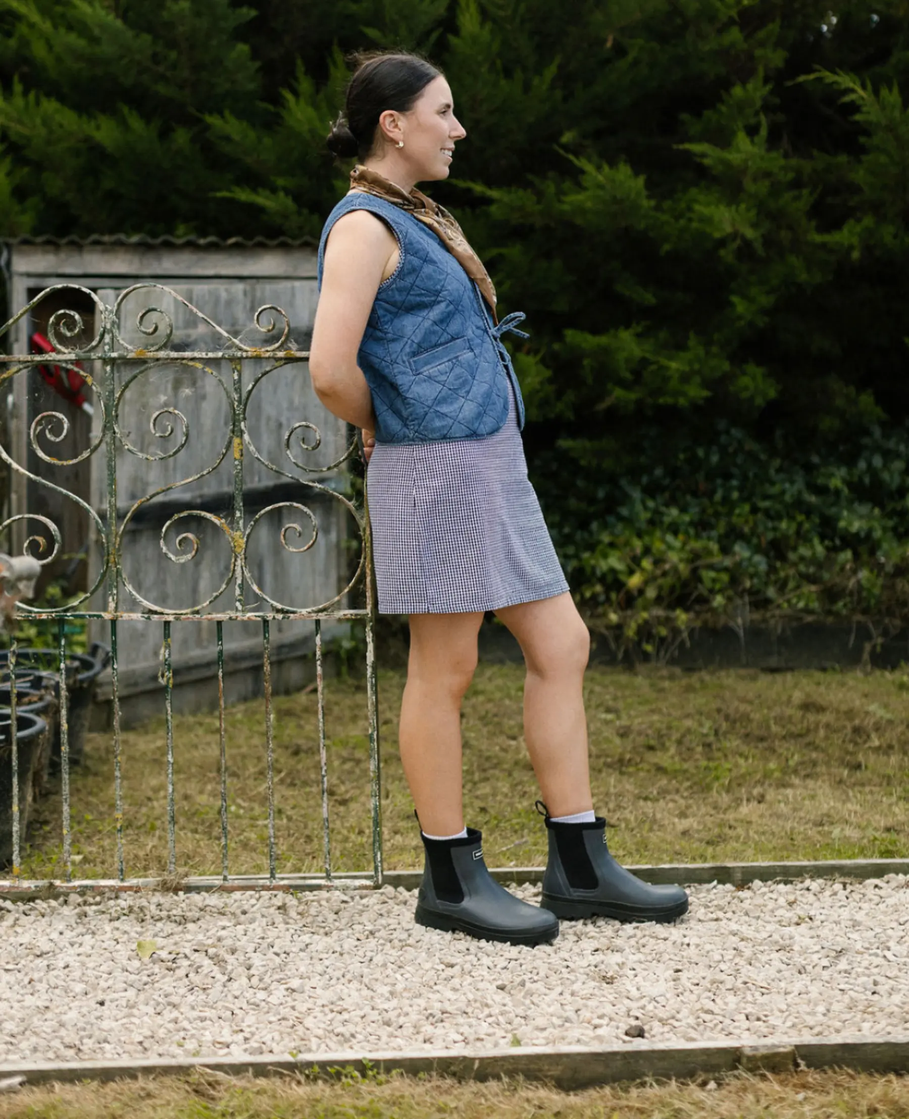A person with long hair in a braid stands outdoors on gravel, wearing a blue quilted vest over a striped dress and black ankle boots. Green bushes and a rustic metal gate are in the background.