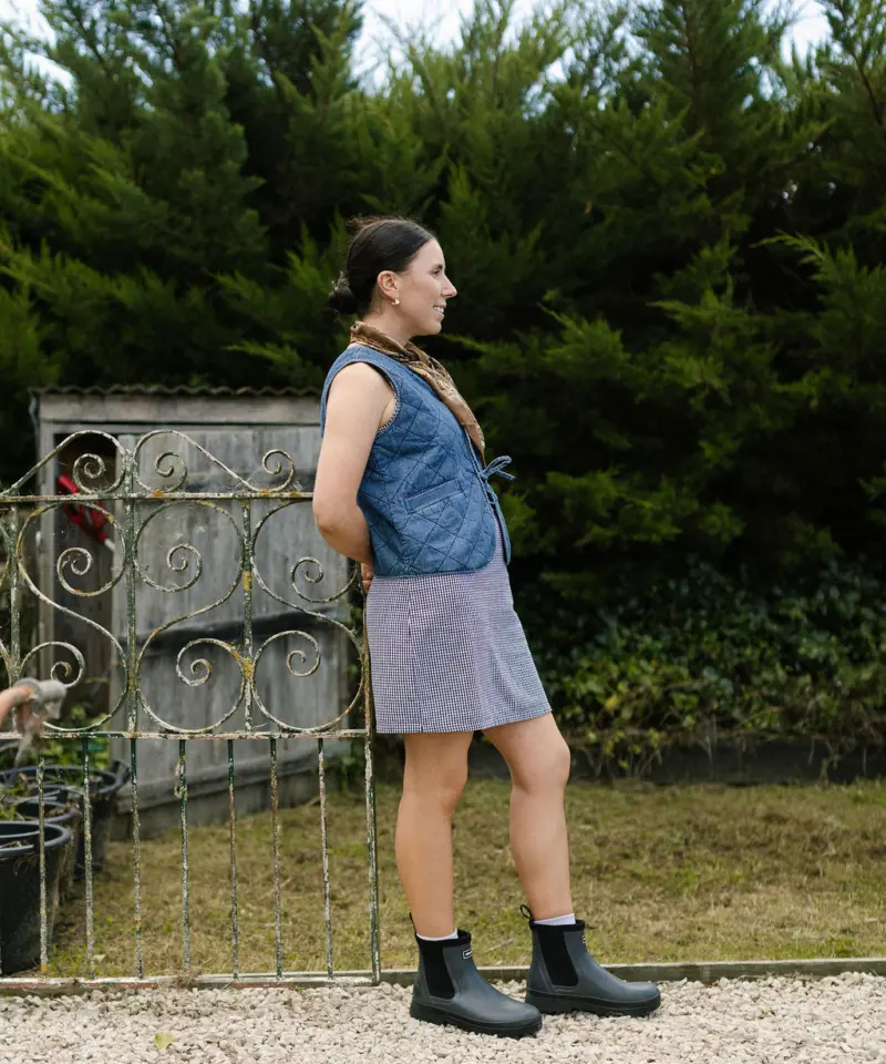 A woman stands outdoors on a gravel path, wearing a blue quilted vest, a short skirt, and black boots. She is facing sideways, with green bushes and a decorative metal gate behind her.