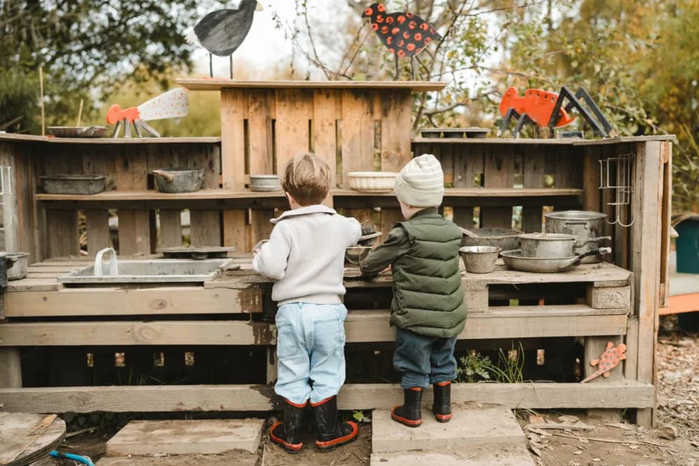 Two young children with light hair, seen from behind, play at a rustic outdoor mud kitchen with metal pots and pans. Homemade animal crafts decorate the wooden structure. The kids wear jackets and rubber boots.