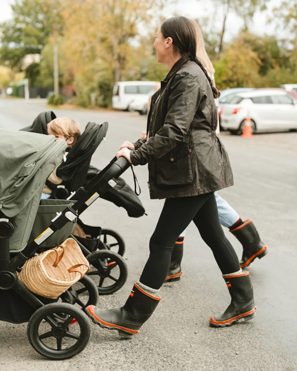 Two women wearing black rain boots and jackets push strollers with children along a wet street. Parked cars and trees with autumn foliage are visible in the background.