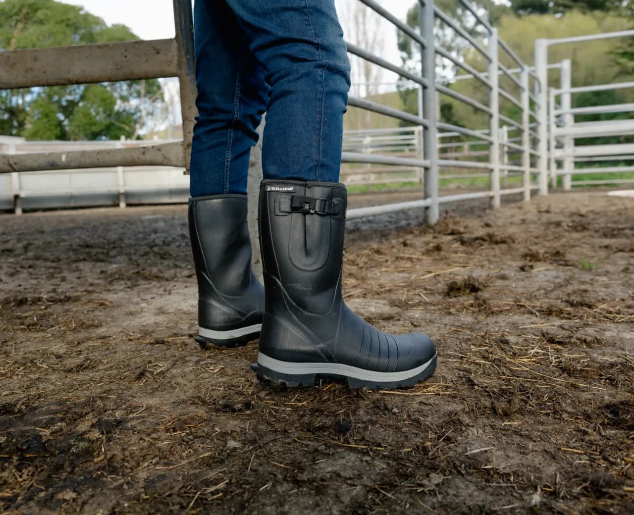 Person wearing black rubber boots and blue jeans stands on muddy ground inside a fenced outdoor area, possibly a farm or animal pen.