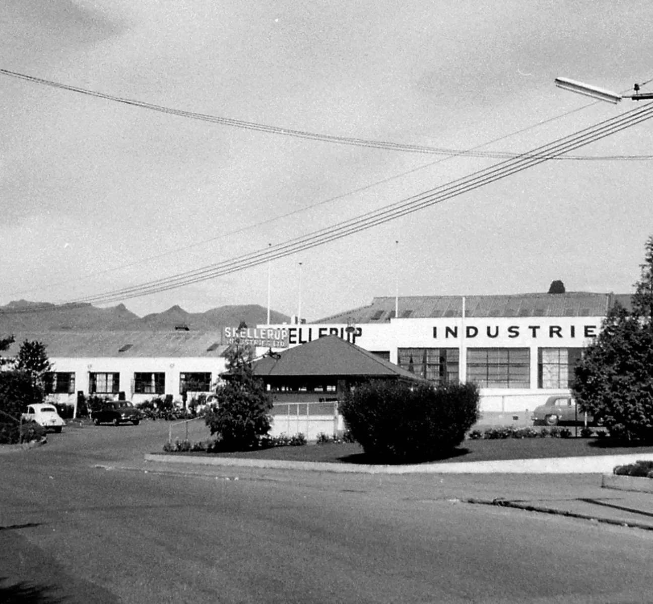 Black and white photo of an industrial building with “SKALLERUP INDUSTRIER” on the facade. Cars are parked outside, and bushes line the street in front. Power lines cross overhead, with mountains visible in the background.