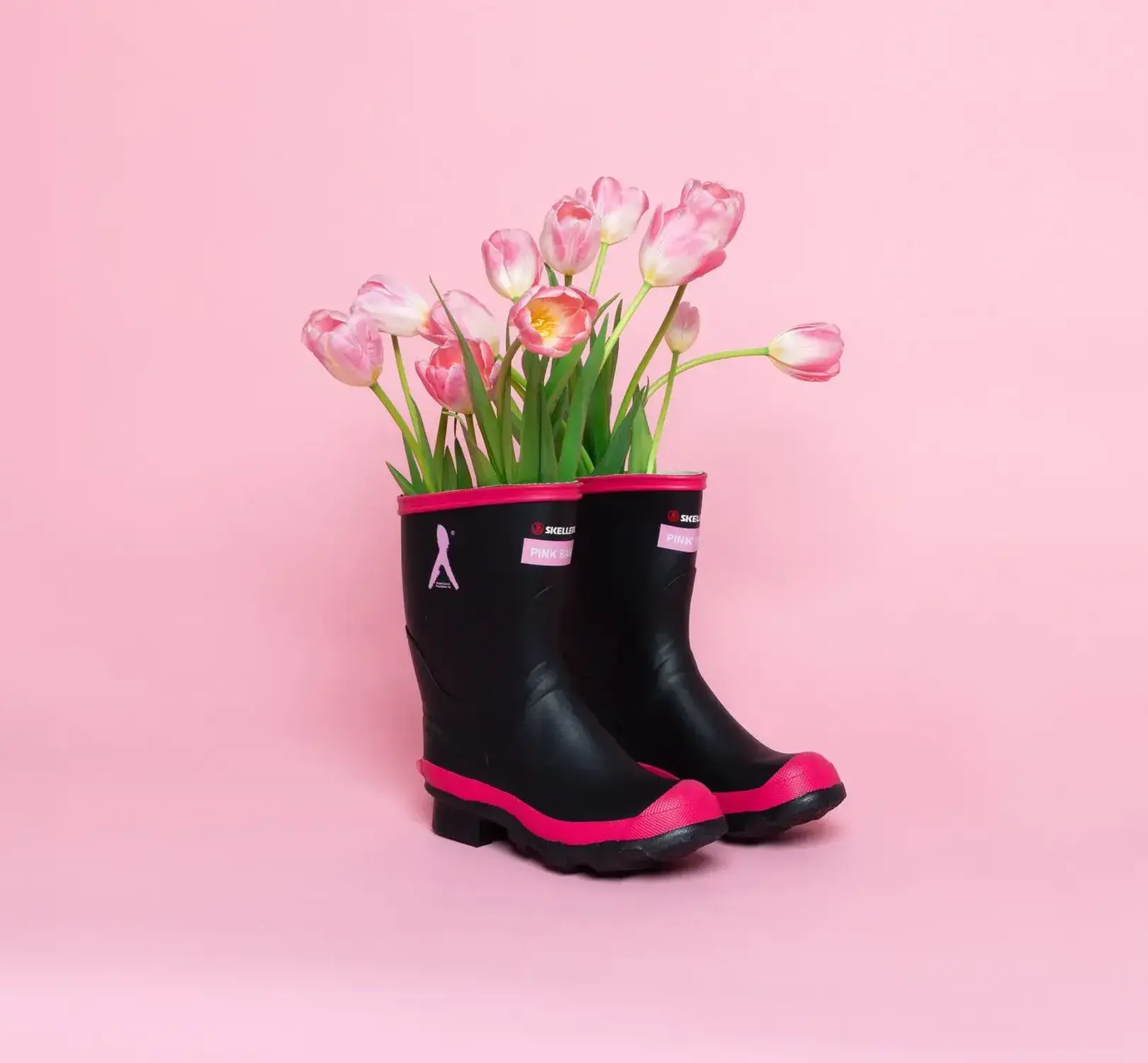 A pair of black rain boots with pink trim are filled with pink tulips and placed against a solid pink background.