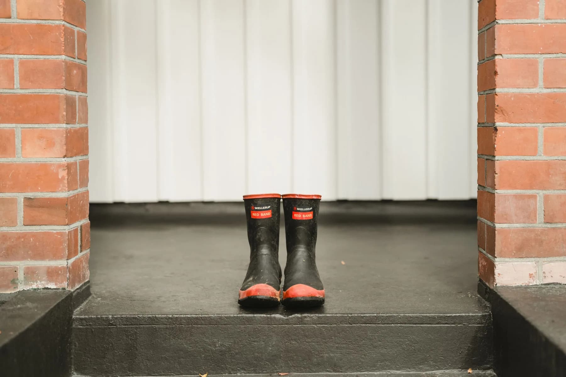 A pair of black rain boots with red soles and trim sits side by side on a black porch step between two brick columns, in front of a white paneled wall.