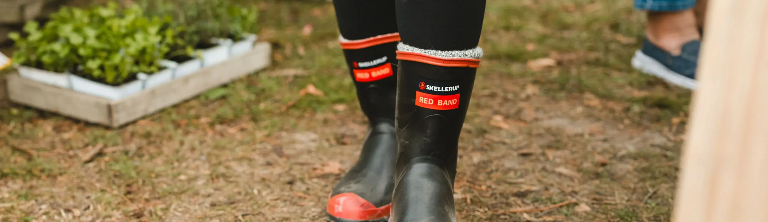 Person wearing black and red rubber boots standing on grass near seedlings in trays; other people with visible legs and shoes are in the background.