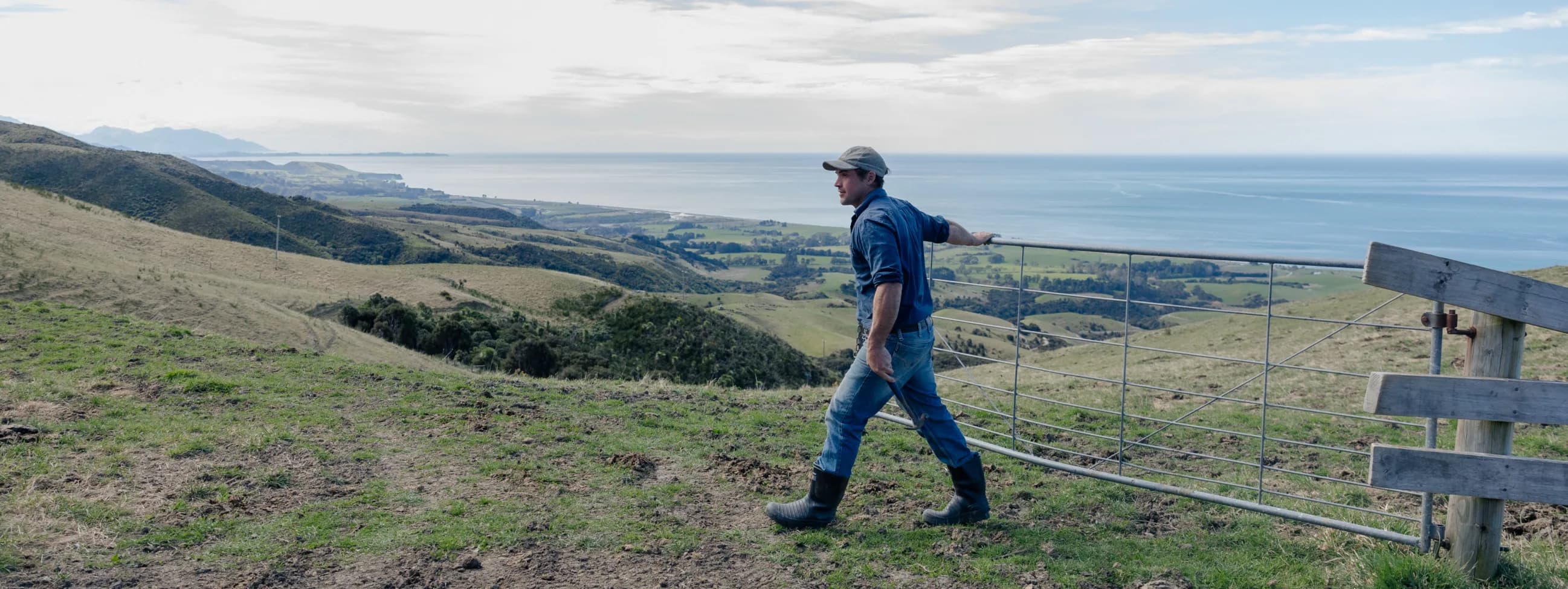 A person wearing jeans, boots, and a cap walks near a metal gate on a grassy hill, with a scenic view of the coastline, ocean, and distant mountains under a cloudy sky.