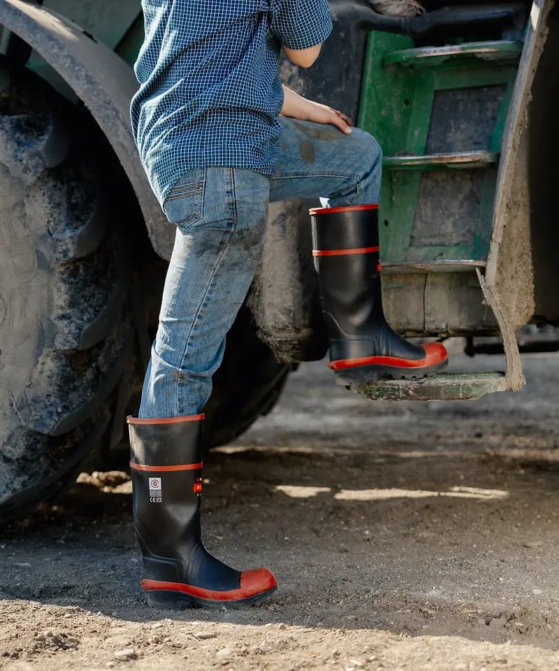 A person wearing blue jeans, a blue checked shirt, and black rubber boots with red soles steps onto a green tractor, with only their lower body visible. Dirt and tire treads are seen on the ground.
