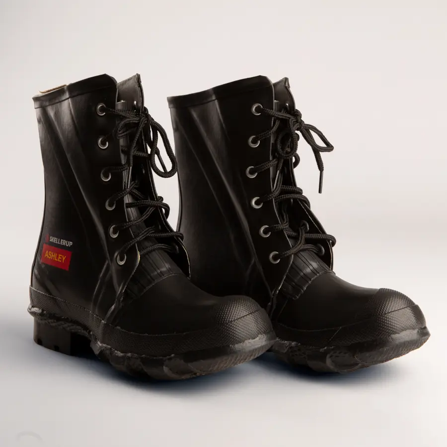 A pair of black work boots with laces sit side by side on a light background. One boot has a red name tag labeled Ashley and Skellerup on the side.