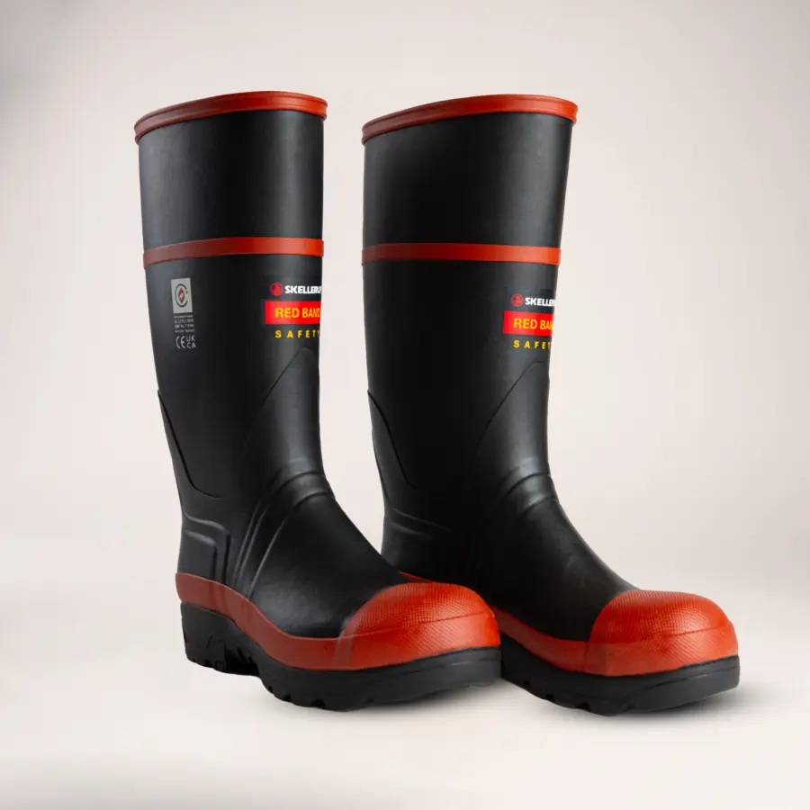 A pair of black rubber safety boots with red accents on the toe, top, and sole, standing upright on a light background. The boots have labels with safety information on the sides.